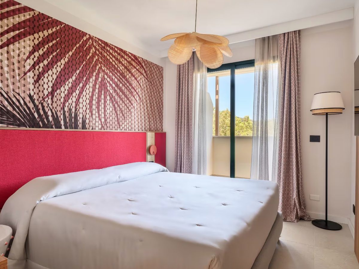 A double bed with a deep pink headboard below a large palm-leaf mural, a woven pendant light and long curtains at the balcony door.