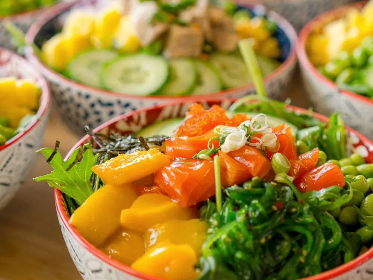 Poke bowls of salmon, mango, cucumber, seaweed salad and edamame in patterned ceramic bowls on a wooden table.