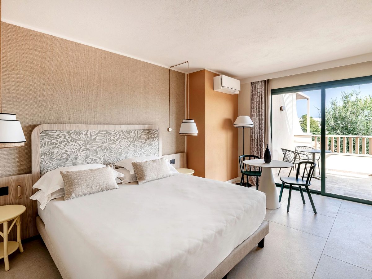 A double bed with a printed headboard against a woven wallcovering, pendant lights either side and sliding doors onto a furnished terrace.