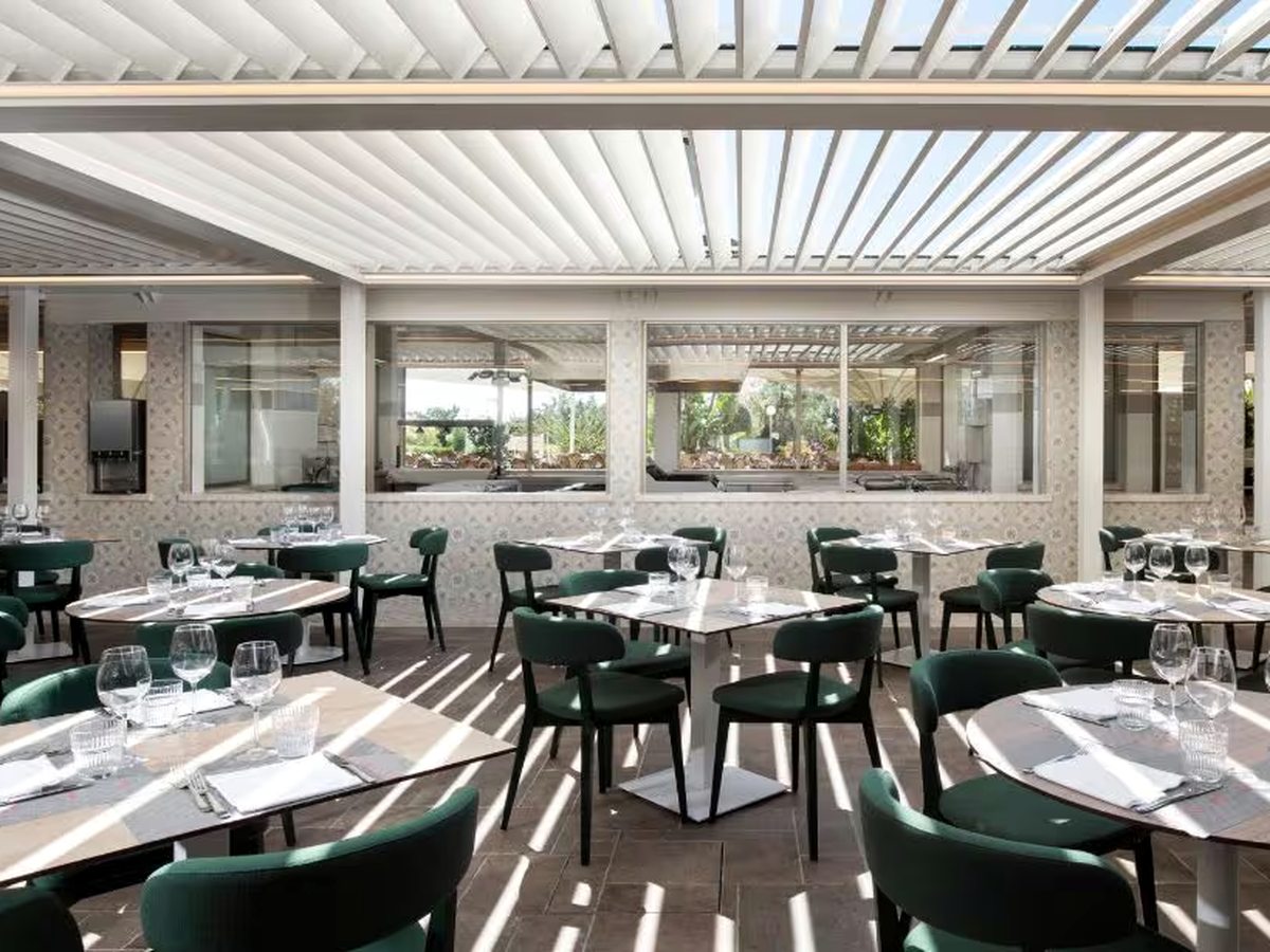 A restaurant terrace under a white louvred roof, with laid tables and dark green upholstered chairs and open sides looking onto the gardens.
