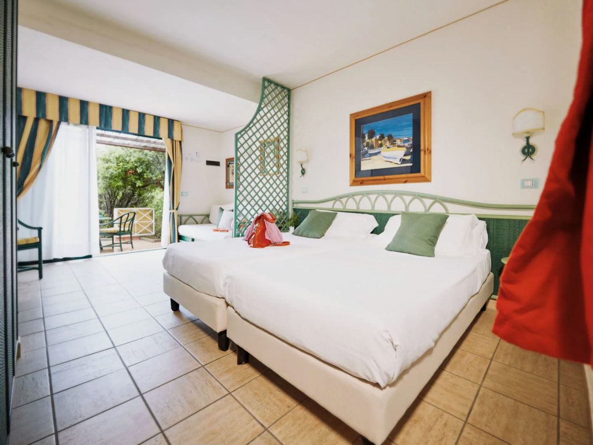 A tiled twin-bedded room with a green rattan headboard and lattice screen, a further bed behind it and doors open onto a shaded garden terrace.