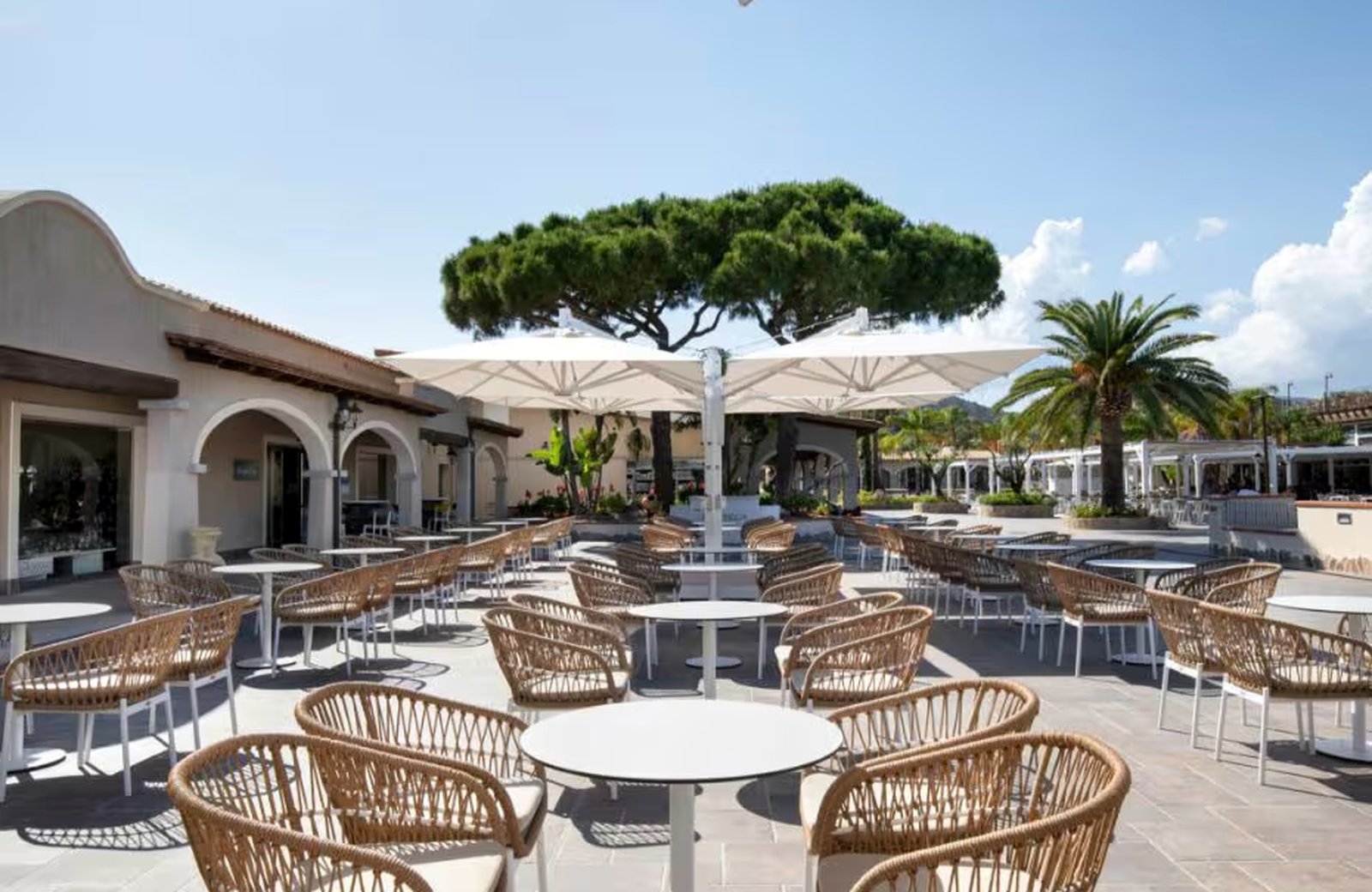 Café tables and woven chairs under white parasols on the central square at VOI Tanka Village, with an arcaded white building along one side and a stone pine and palms behind.