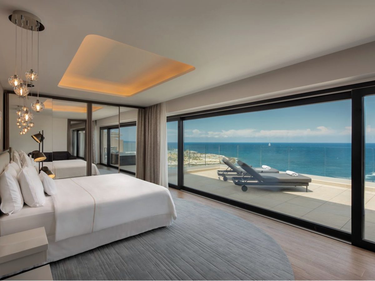 A suite bedroom with a king bed and cluster pendant lights, with a full-width sliding glass wall onto a terrace with sun loungers and the open sea beyond.