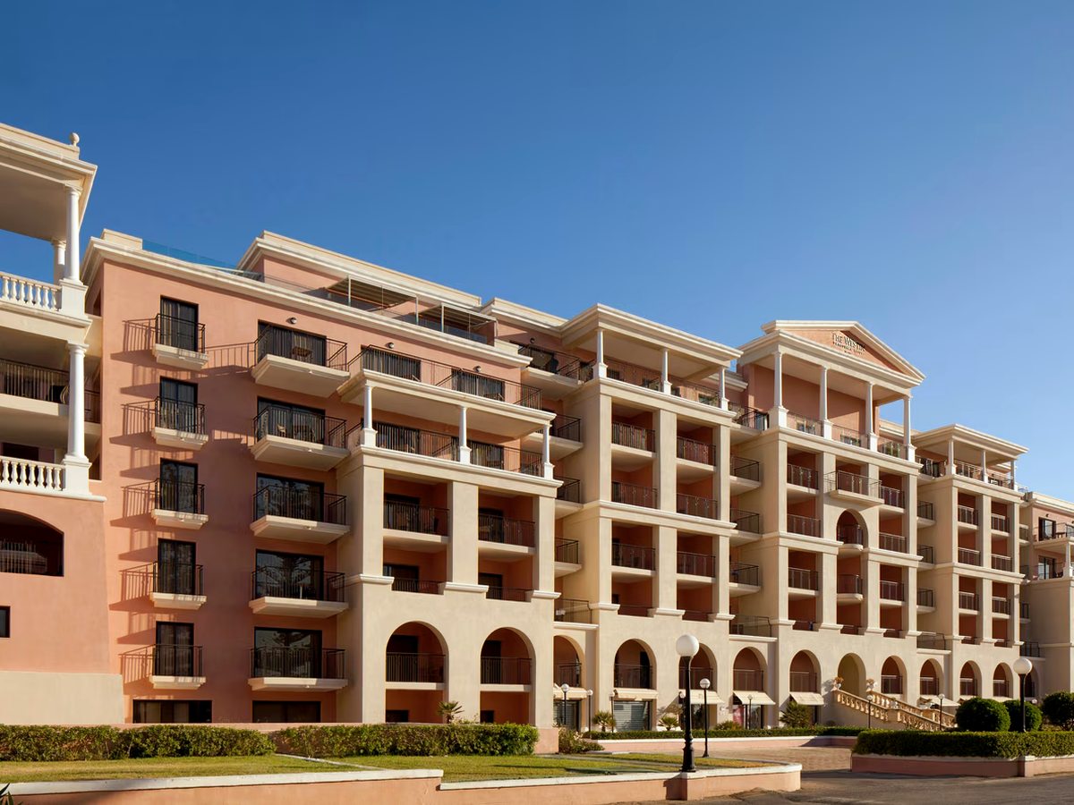 A long pink and cream hotel block of six storeys with rows of balconies and arched ground-floor openings under a clear blue sky.