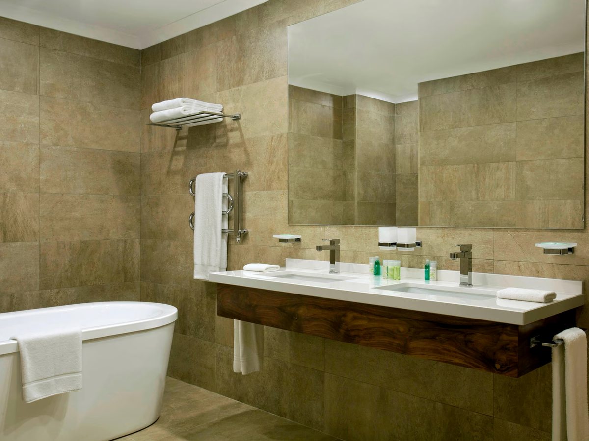 A bathroom clad in beige stone tiles with a freestanding oval bath, a wide twin-basin vanity in dark wood and a large mirror.