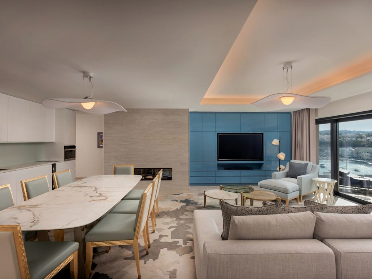 A suite living and dining area with a long marble table and pale green chairs, a kitchen run with an oven, a fireplace and a blue media wall with a television above a corner sofa.