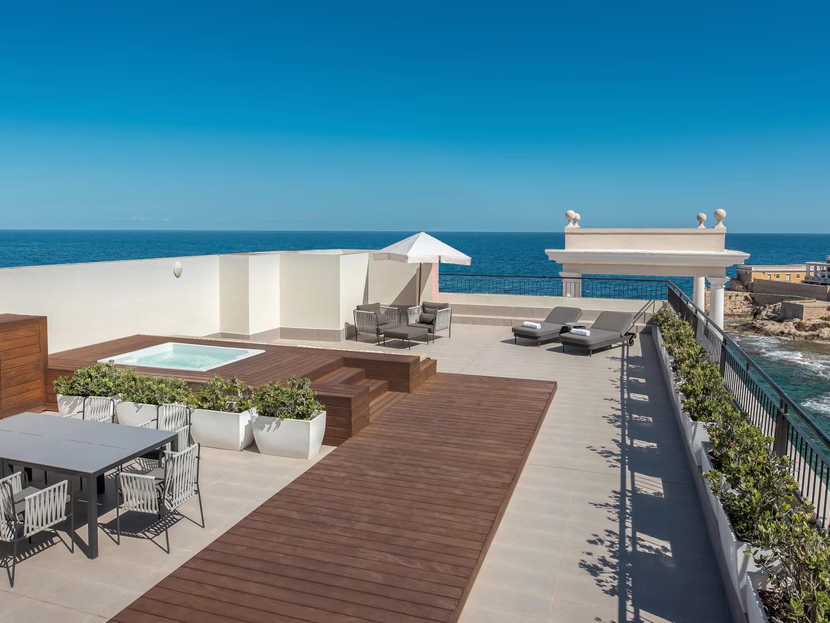 A large suite terrace with a hot tub set into a timber deck, a dining table, sun loungers and a white parasol, with the sea beyond the railings.