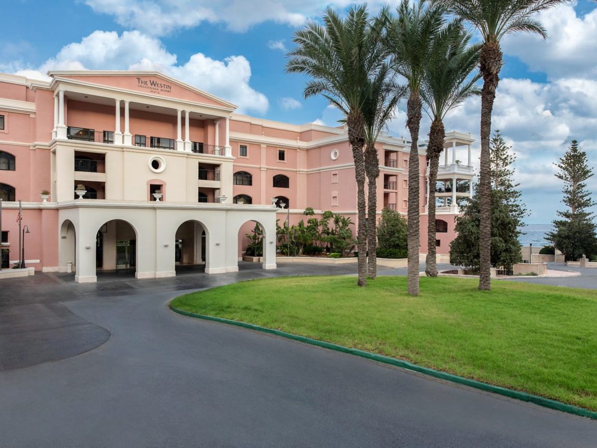 The pink and cream main building behind a white arched entrance portico, with palms, a lawn and a curved drive in front.
