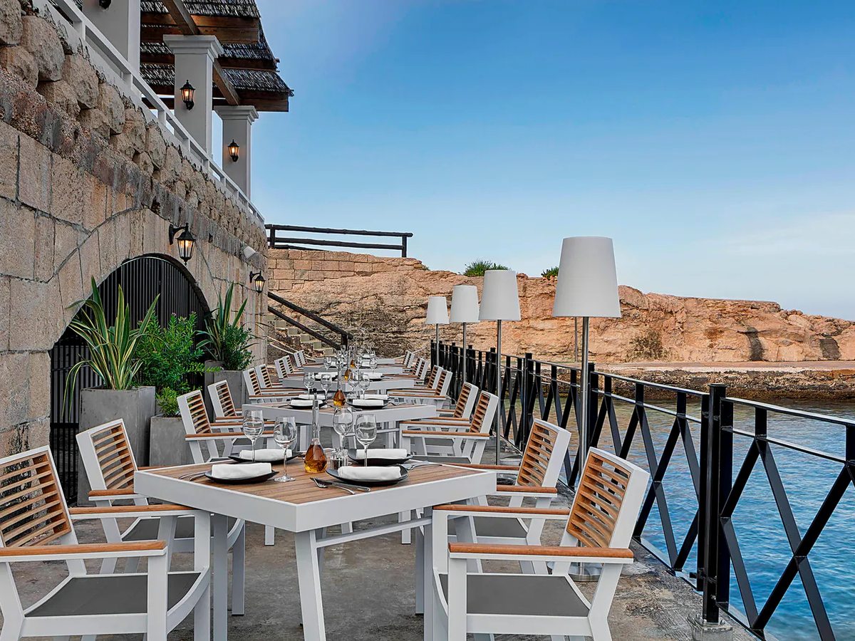 Tables laid with wine glasses on a narrow waterside terrace between a stone wall and a black railing, with white floor lamps and low limestone rocks across the water.