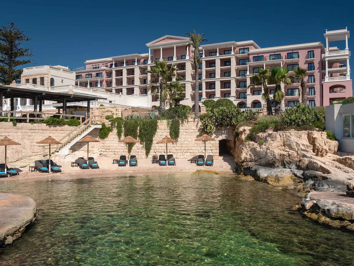 A small sandy cove with clear shallow green water, thatched parasols and loungers on the sand below the pink hotel building and its terraces.
