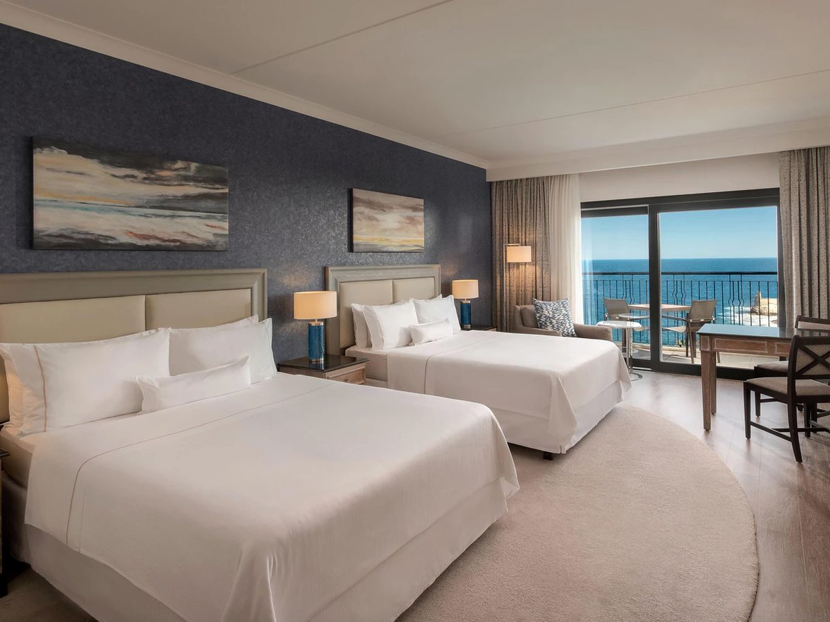 A hotel room with two double beds against a dark blue wall, seascape paintings above them, a desk and sliding doors onto a balcony over the sea.