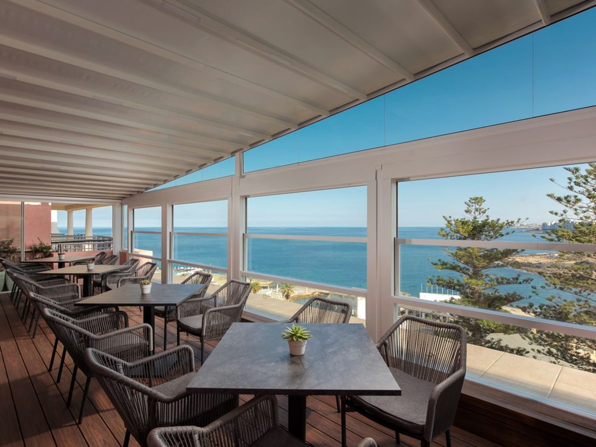 A glazed dining terrace with dark tables and woven chairs on a timber floor, with wide windows onto the blue sea and the coastline.