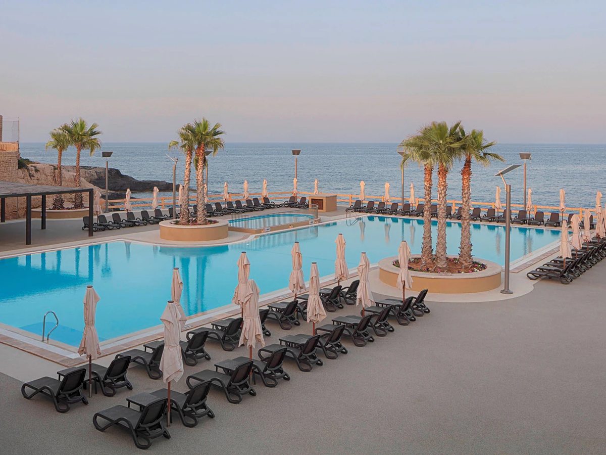 A large rectangular pool at sea level with palms in raised planters, rows of loungers and parasols on the deck and a smaller shallow pool at the far end.