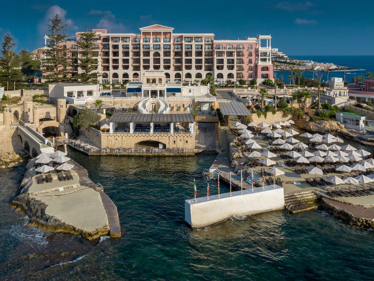 Sea-level terraces of white parasols and loungers on the rocks around a small inlet, with a white swimming platform and the pink hotel building above.