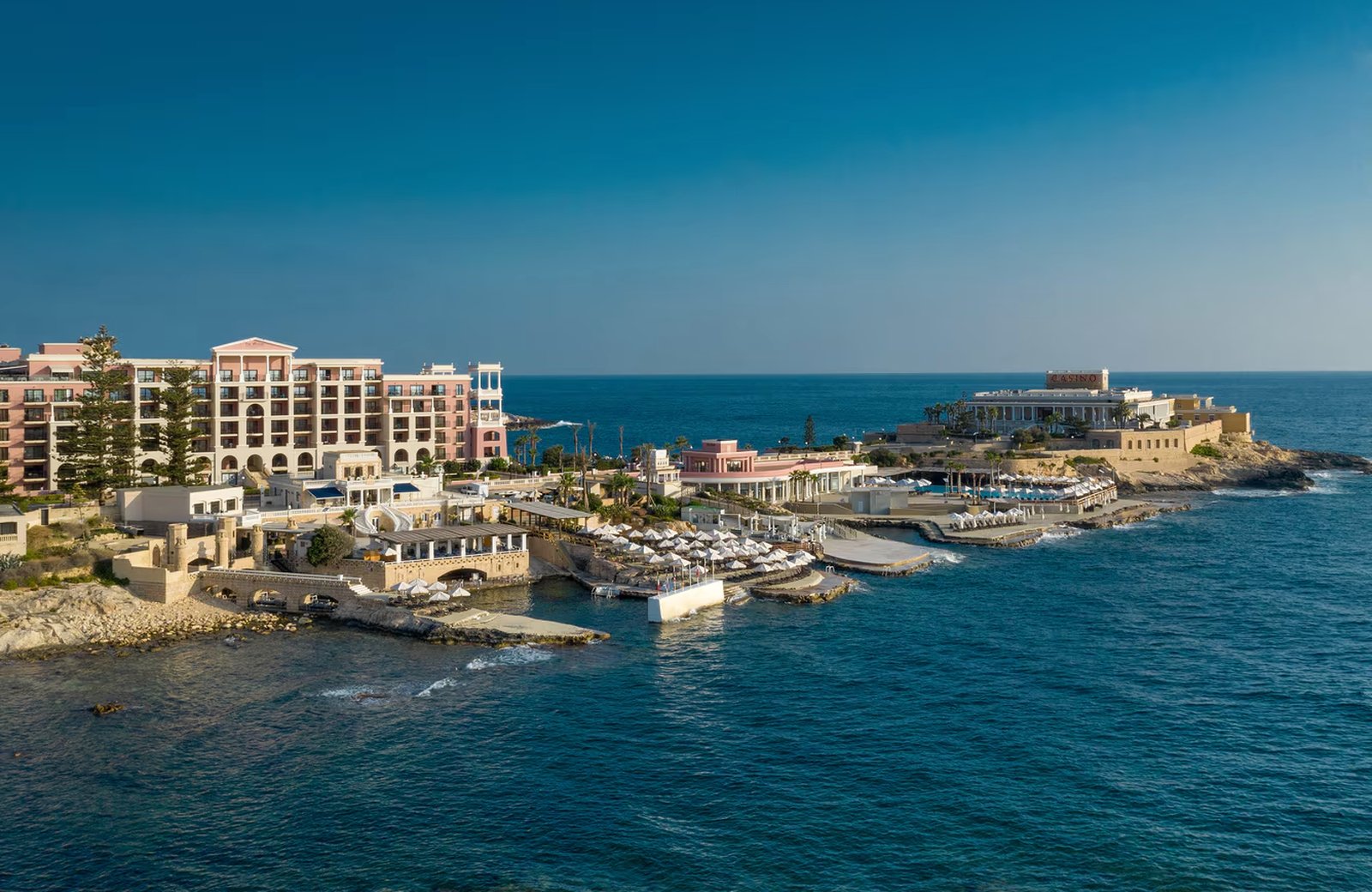The Westin Dragonara Resort seen from the sea: the pink main building above sea-level terraces of white parasols and swimming platforms, on a rocky peninsula with the casino on the headland beyond.