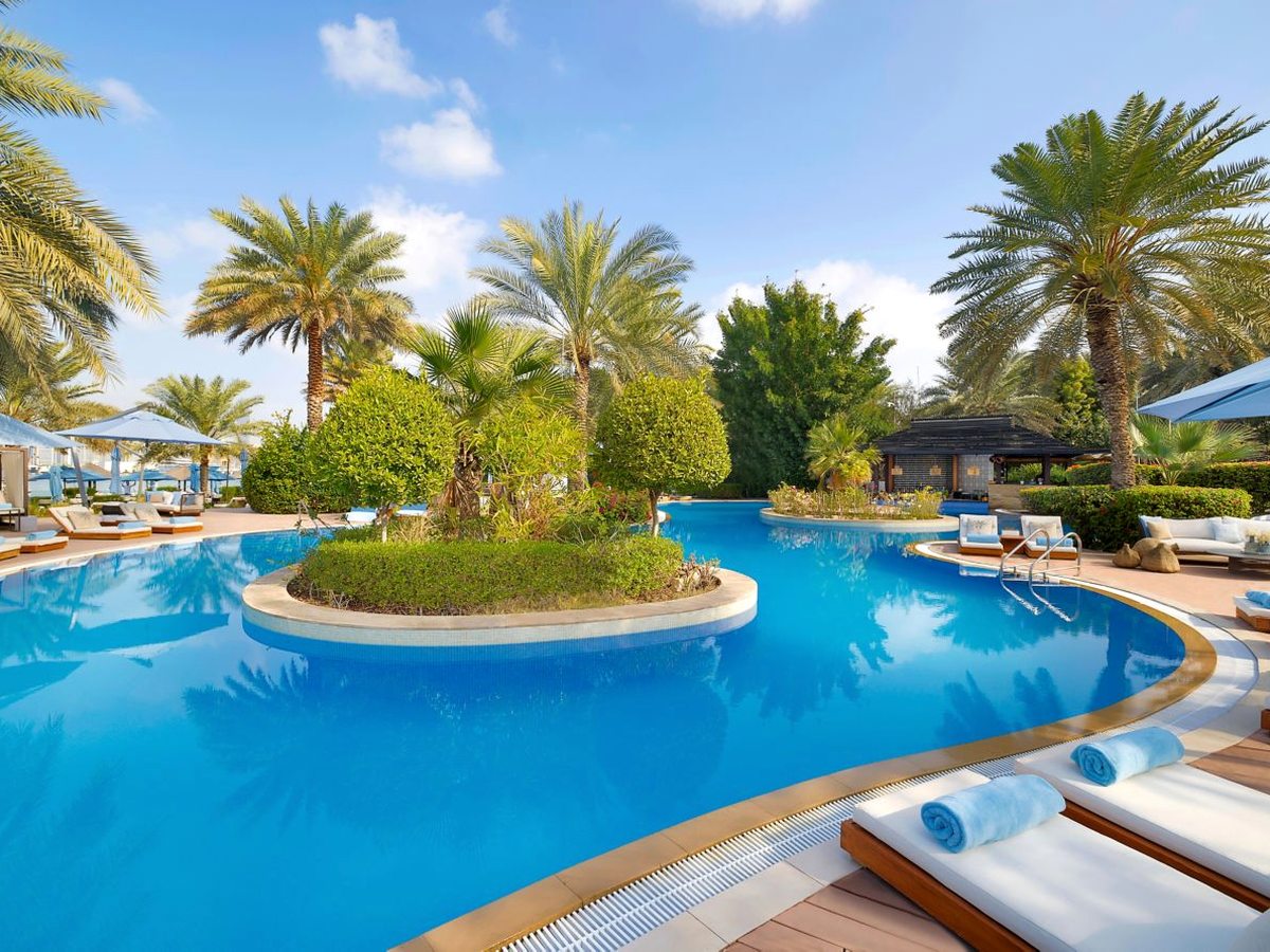 A curving blue swimming pool wrapped around a planted island of shrubs and palms, with white sun loungers and blue towels on the paved surround.