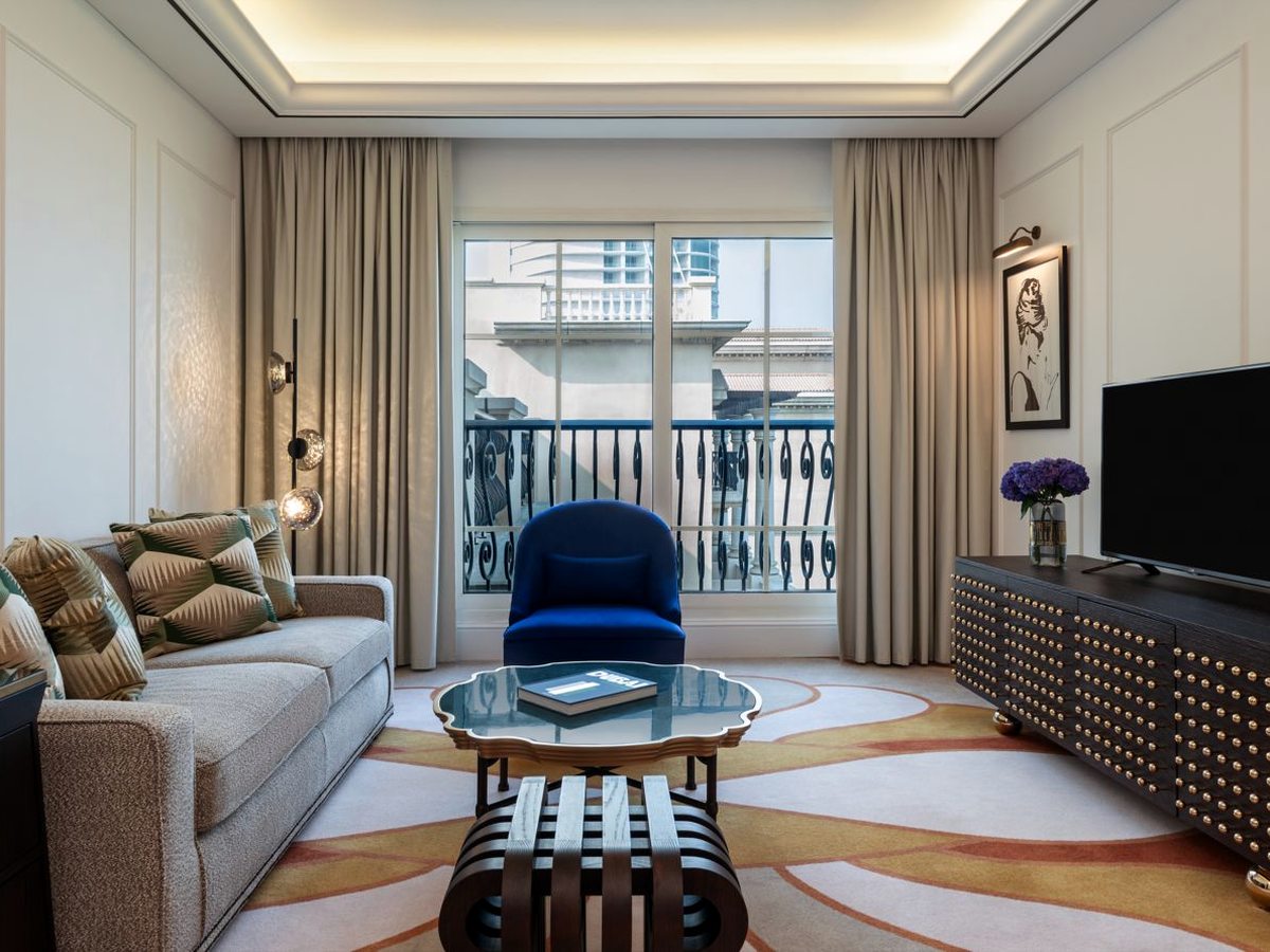 A suite sitting room with a pale grey sofa and cushions, a royal blue armchair by tall balcony doors, a scalloped glass coffee table on a patterned cream and ochre rug, and a studded dark sideboard with a television.