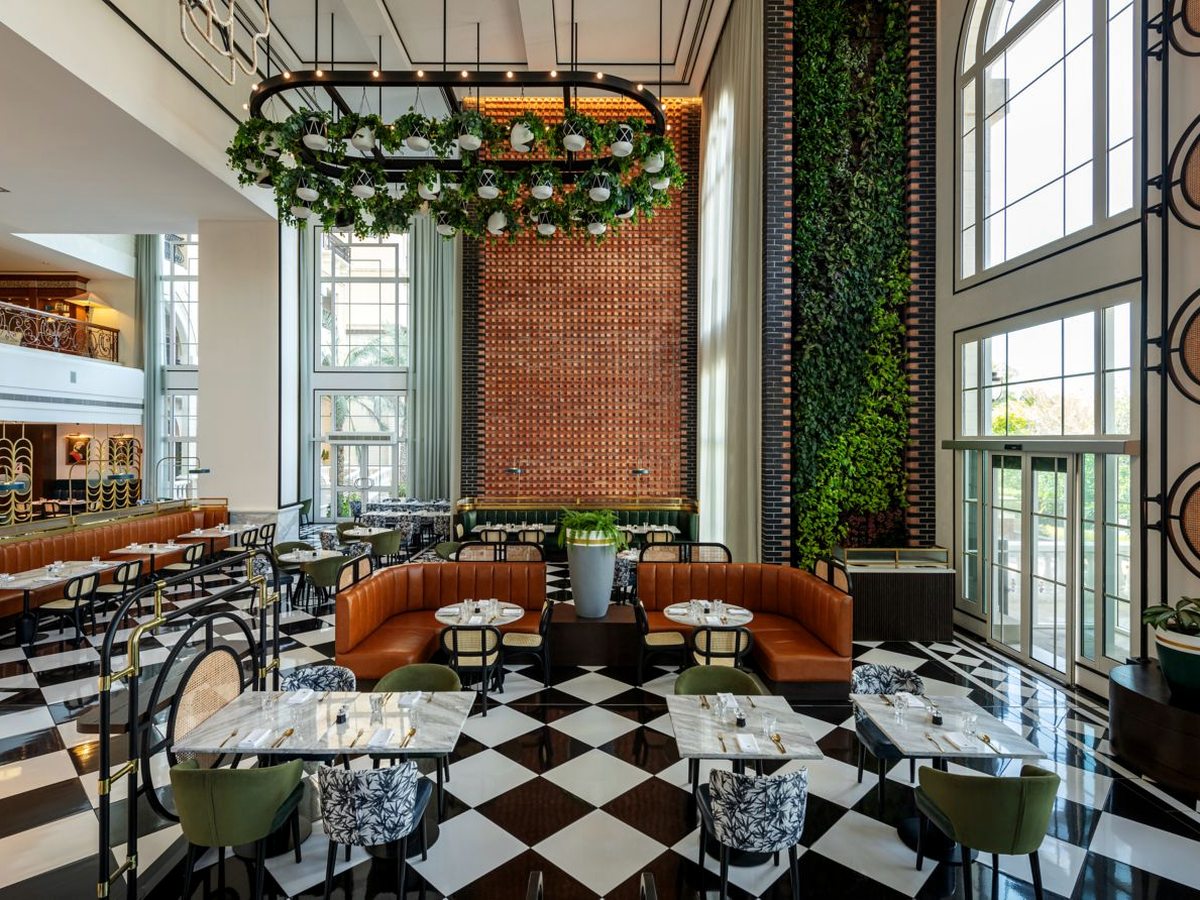 A tall dining room with a black and white chequered marble floor, tan leather banquettes, green velvet chairs and marble tables, a brick feature wall, a wall of trailing plants and a circular greenery-hung light fitting.