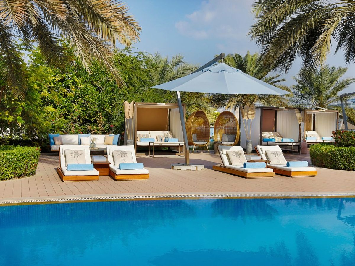 A row of timber-framed poolside daybeds with white curtains and cream cushions, a large parasol and hanging egg chairs on a decked terrace beside a blue pool.