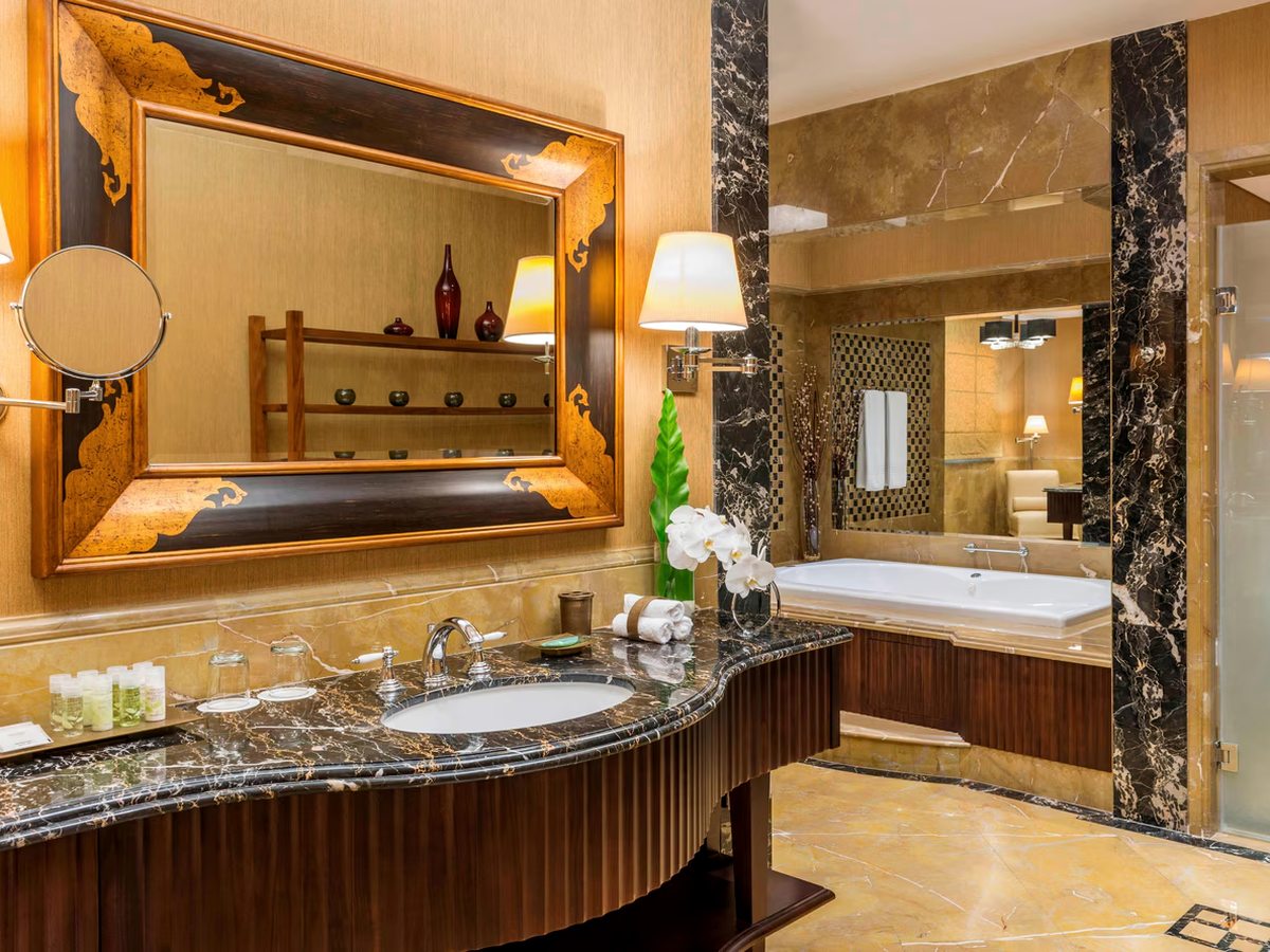 A bathroom with a dark marble vanity and single basin below a wide inlaid wooden mirror and wall lamps, with a deep white tub set into a marble surround and a glass shower enclosure beyond.