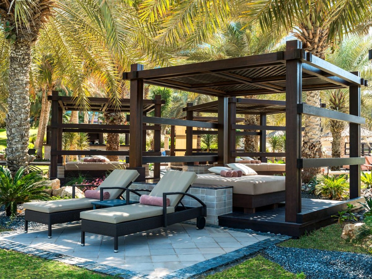 A dark timber cabana with a slatted roof sheltering a wide cushioned daybed, with two padded sun loungers on a paved terrace in front and date palms and shrubs all around.