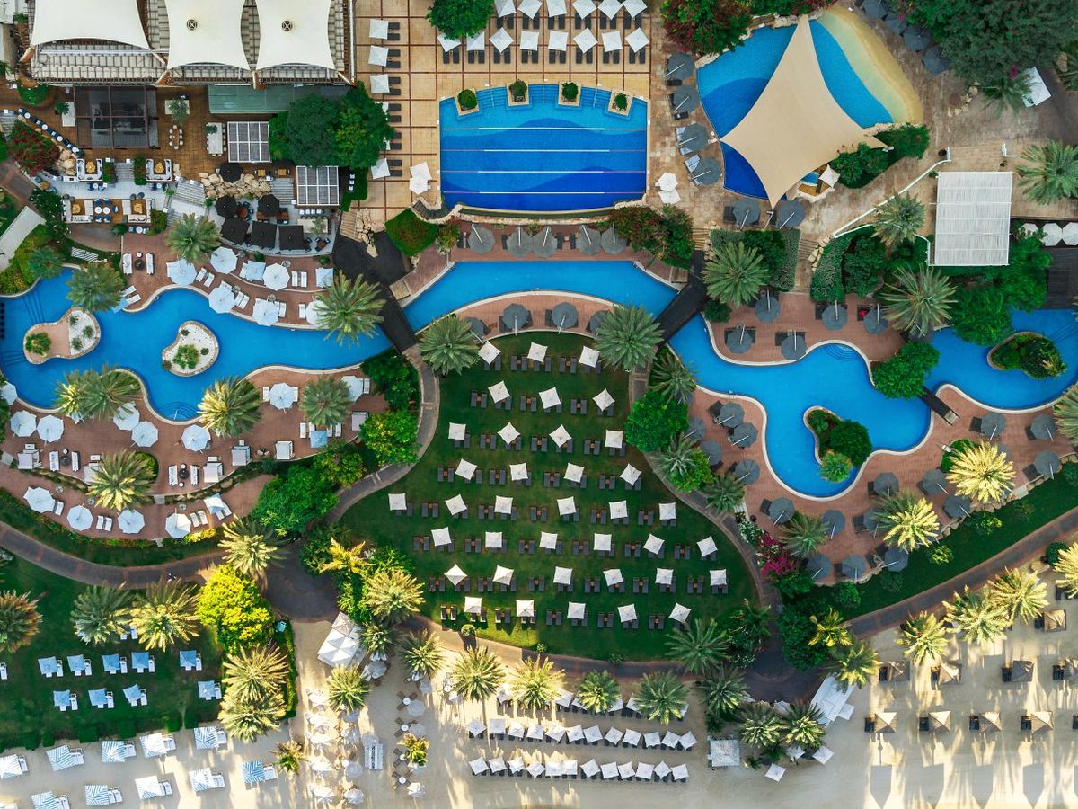 Freeform blue pools and a rectangular lap pool set among lawns, palm gardens and ranks of white parasols and sun loungers within the resort grounds.