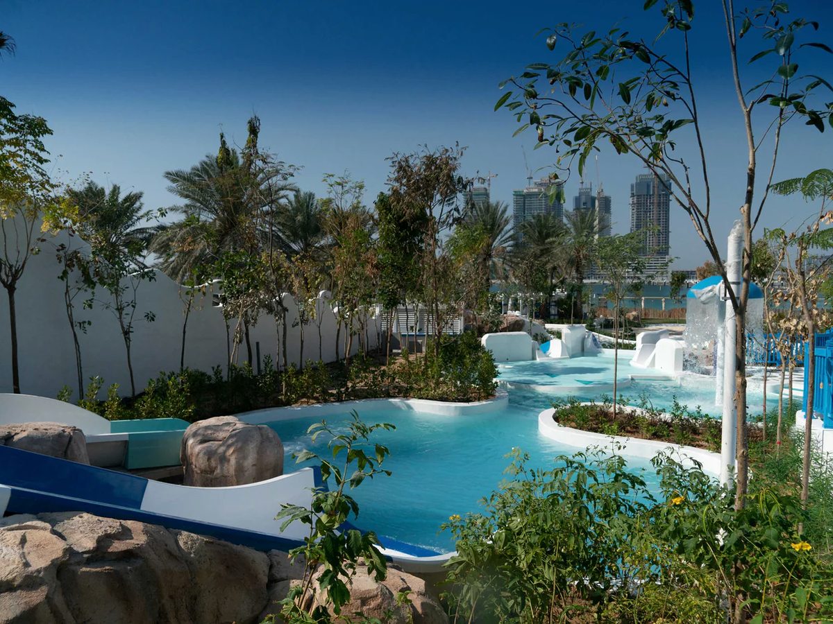 A winding turquoise water channel running between white rendered walls and rock features, with young trees and palms planted along it and high-rise towers on the skyline.