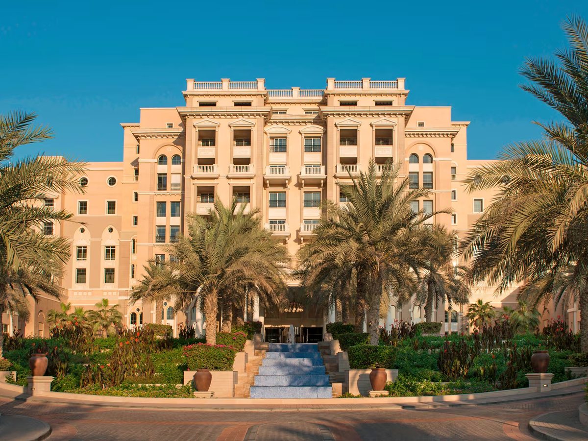 A large beige neoclassical hotel building with balconied windows and a pedimented centre, framed by tall date palms above a planted circular driveway with a stepped blue water cascade leading to the doors.