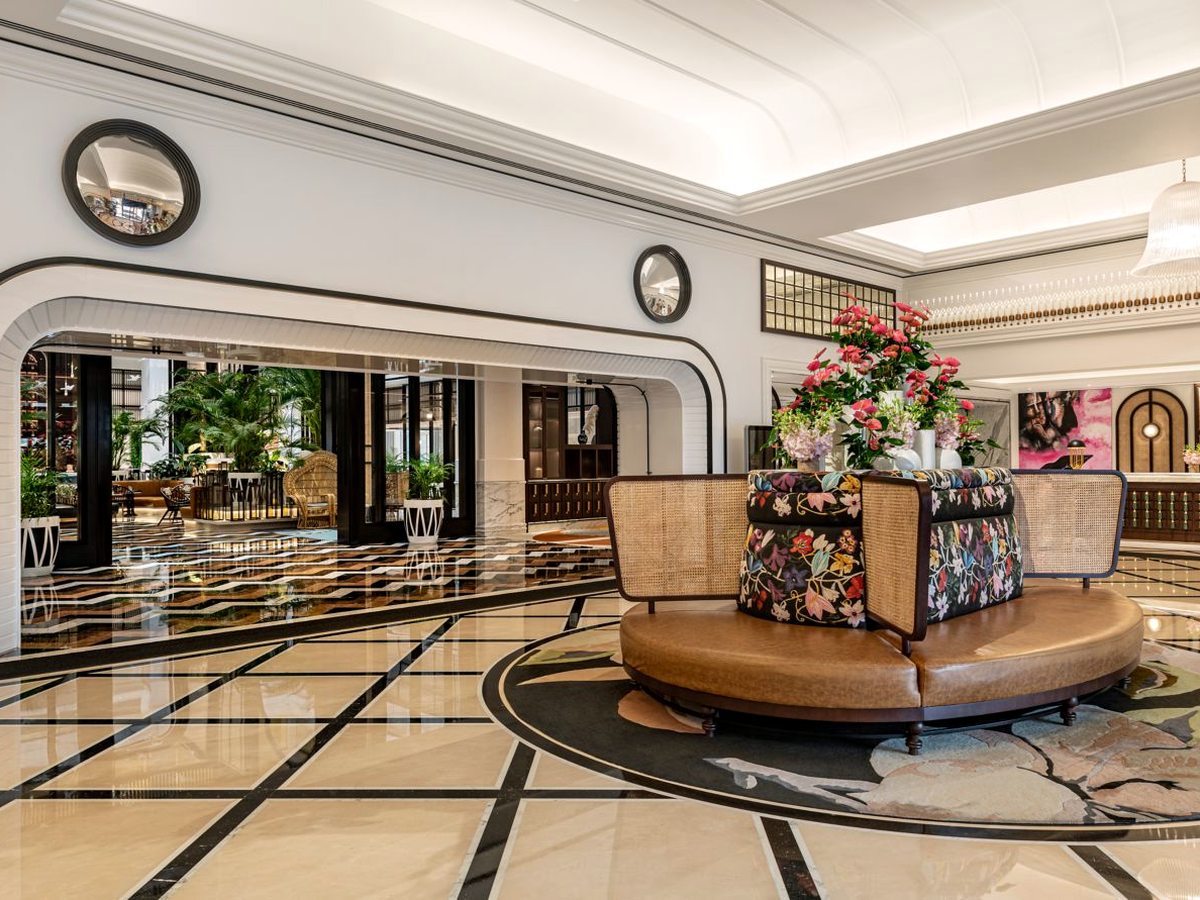 A bright marble-floored hotel lobby with black inlaid lines, a round tan leather banquette topped by a tall pink and white flower arrangement, convex mirrors on white walls and an arched opening through to a planted lounge.