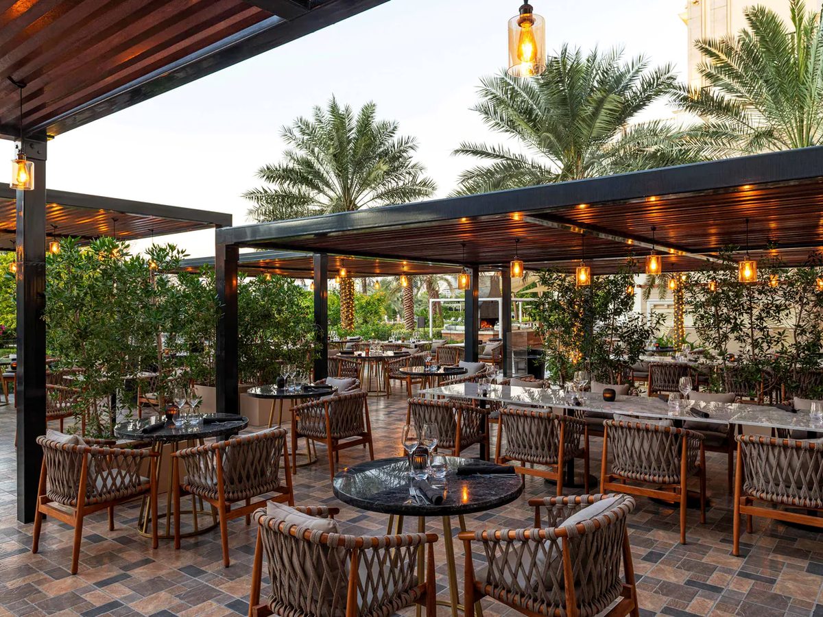 An outdoor restaurant terrace under dark timber pergolas strung with warm pendant lanterns, with woven rope armchairs at black marble tables, planted screens of small trees and date palms behind.