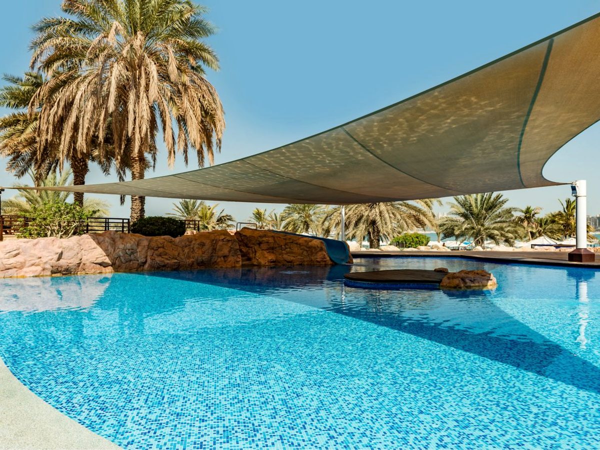 A shallow blue mosaic-tiled pool under a large cream shade sail, with a short slide running down a low rock outcrop into the water and date palms behind.