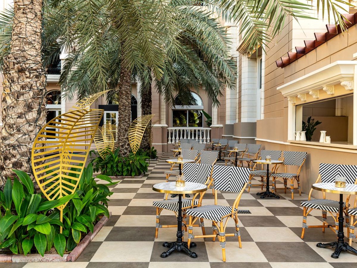 An outdoor café on a beige and cream chequered floor with small marble bistro tables and black and white rattan chairs, yellow leaf sculptures in the planting and date palms in front of a cream stone facade.