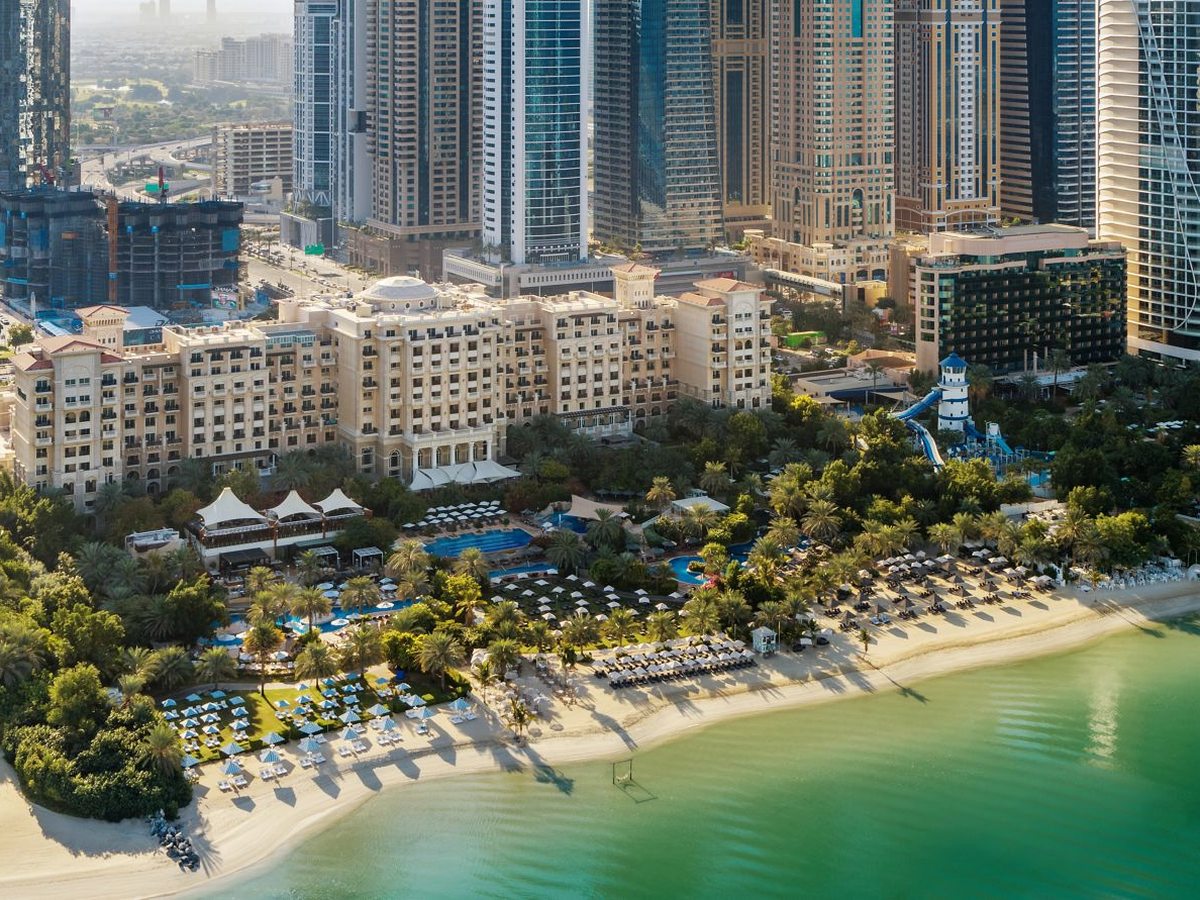 A long pale sand beach lined with rows of sun loungers and parasols, backed by palm gardens, pools and the resort building, with green shallow sea in the foreground and Dubai high-rises behind.