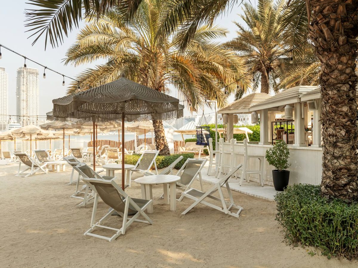 A small white beach kiosk with bar stools at its counter, with white deckchairs and low tables on the sand under fringed and thatched parasols, date palms overhead and towers across the water.