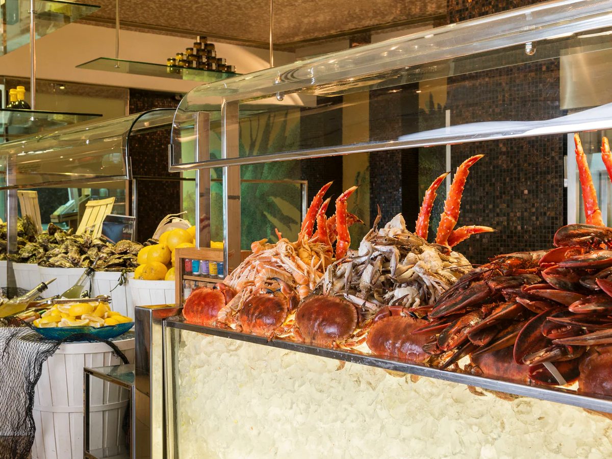 A buffet counter under a curved glass guard, piled with crab claws, crab shells and lobster on crushed ice, with baskets of oysters and bowls of lemons alongside.