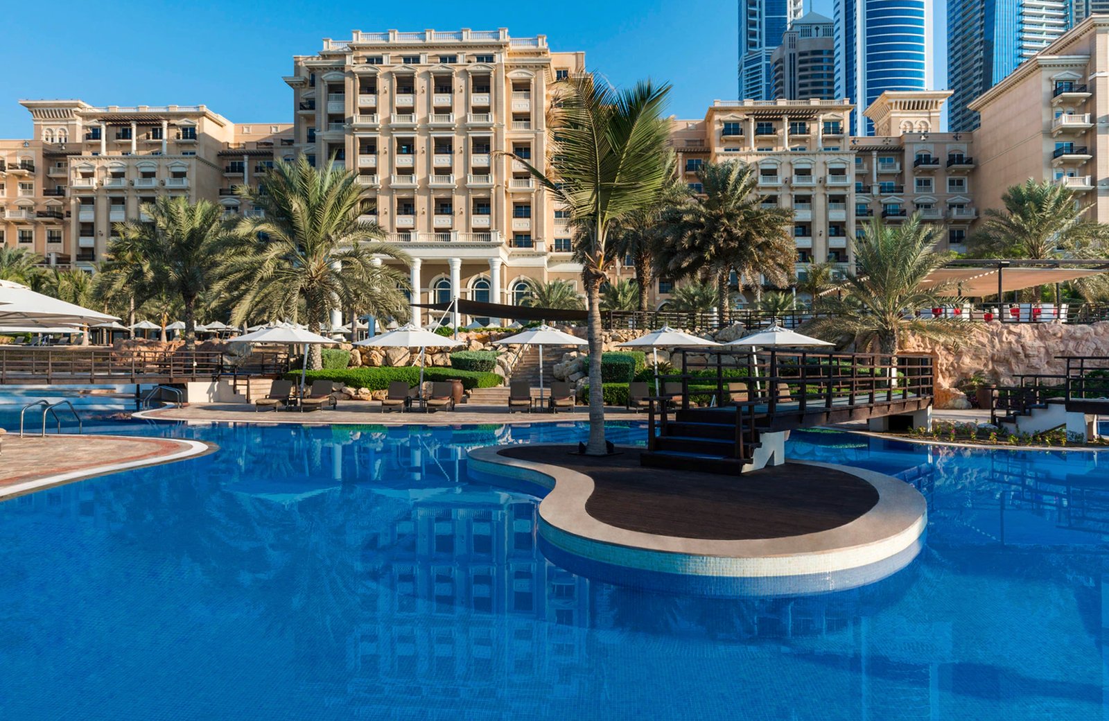 The Westin Dubai Mina Seyahi: a beige neoclassical resort building behind a wide blue pool with a planted deck island, date palms and sun umbrellas, and Dubai Marina towers to one side.
