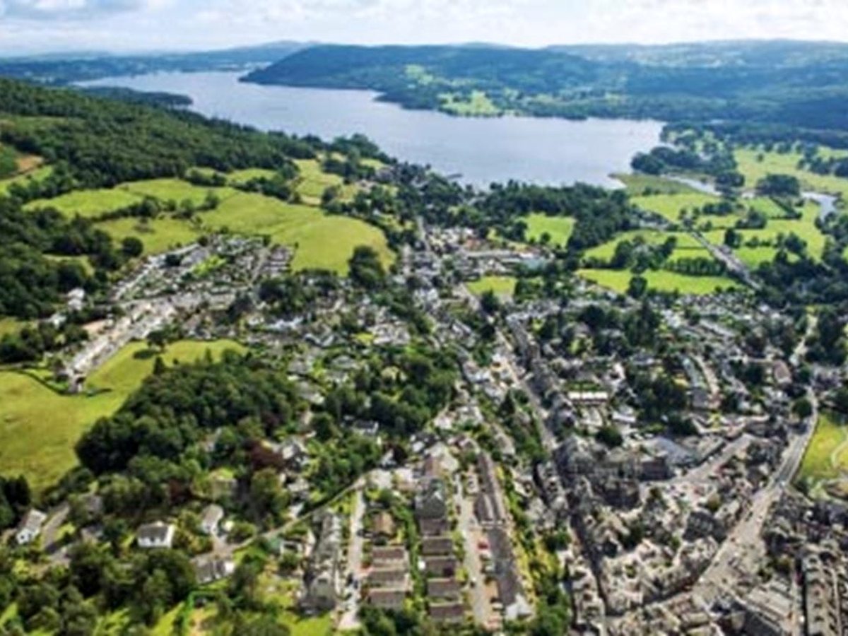 An aerial view of Ambleside spread along the valley floor among green fields, with the river winding into the head of Windermere and wooded hills beyond.