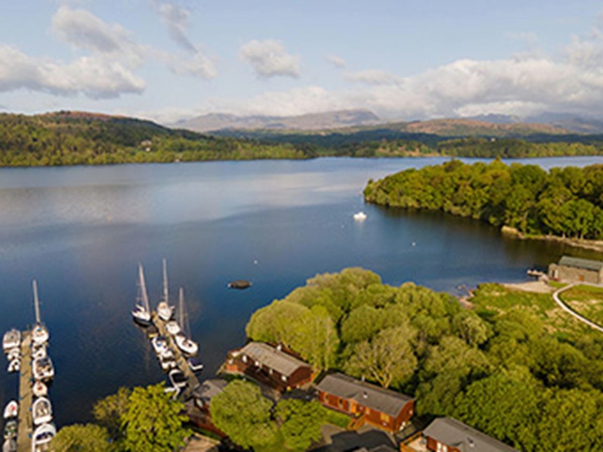 An aerial view of the park's marina with moored yachts, timber lodges among the trees along the shore and Lake Windermere beyond, with wooded hills on the far side under a blue sky.