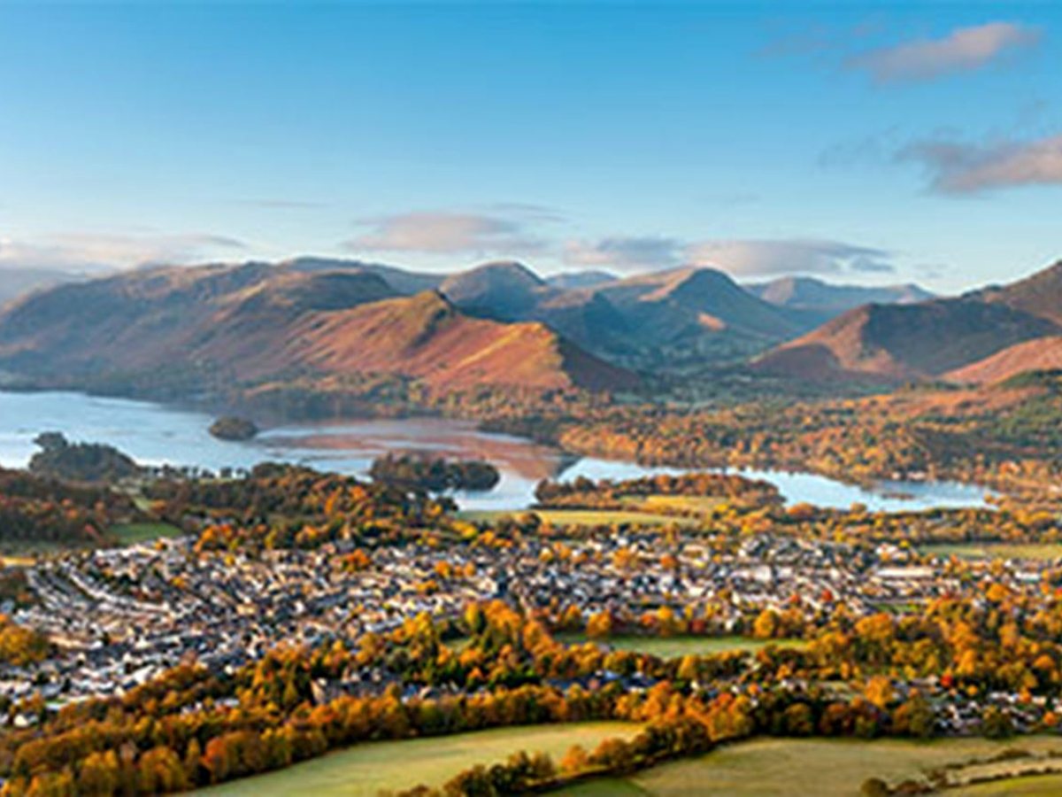 Keswick town below autumn-gold woods and fields, with Derwentwater and its wooded islands beyond and a line of high fells in low sunlight.