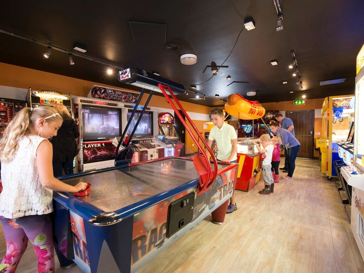 Two children playing at an air hockey table in the arcade, with lit arcade cabinets along the walls and other families at a prize machine behind them.
