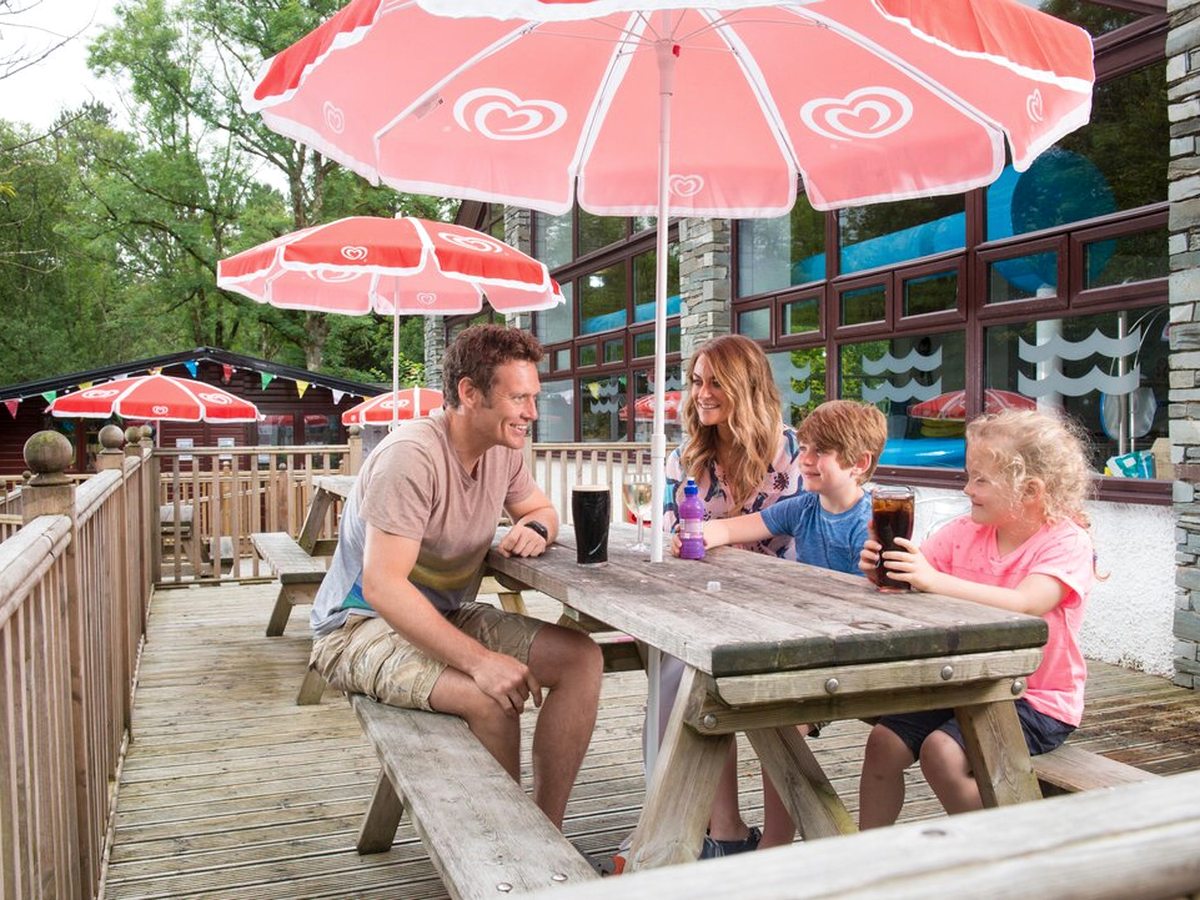 A family of four sitting with drinks at a wooden picnic table on a raised timber deck under red and white parasols, with a glass-fronted building behind them.