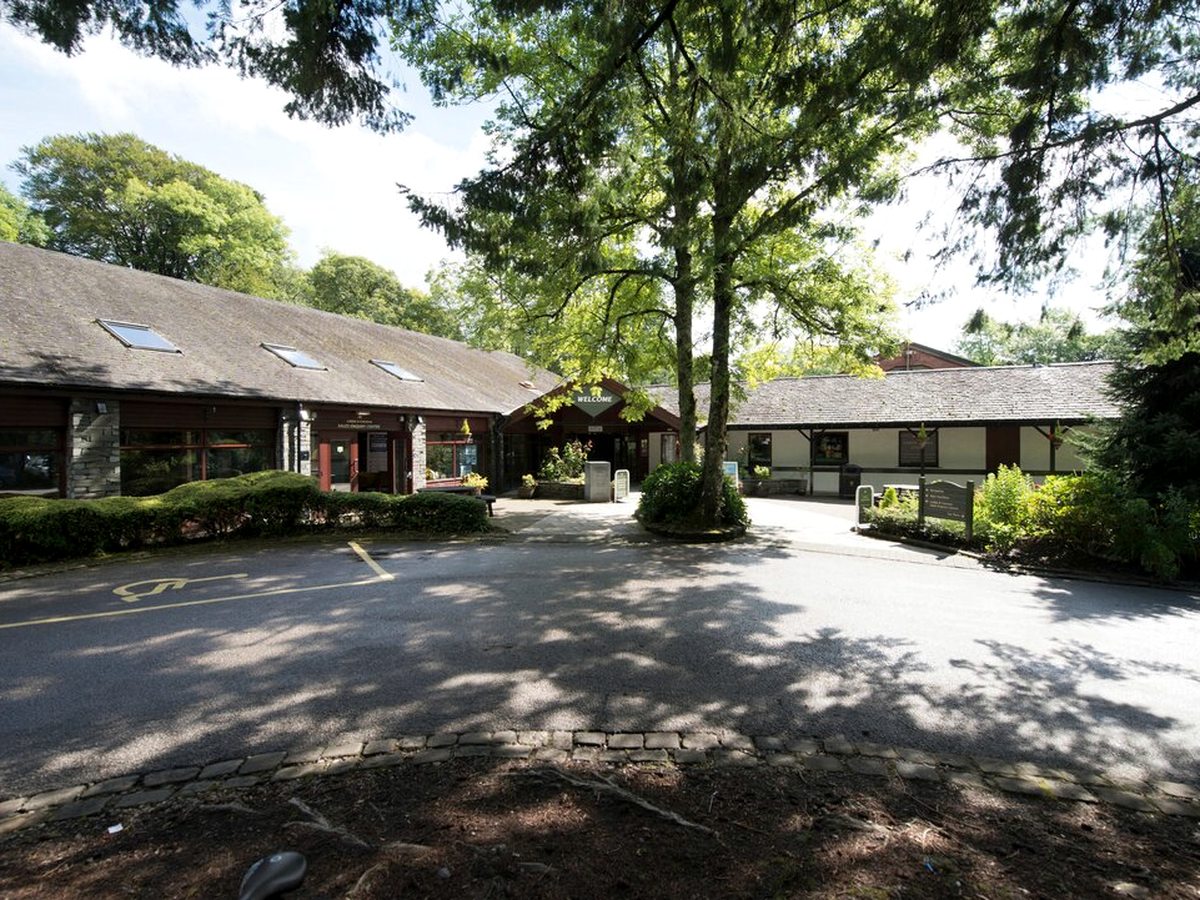 A long, low slate-roofed building of timber and stone with a covered Welcome entrance, standing among tall trees beside a tarmac drive and parking bays.