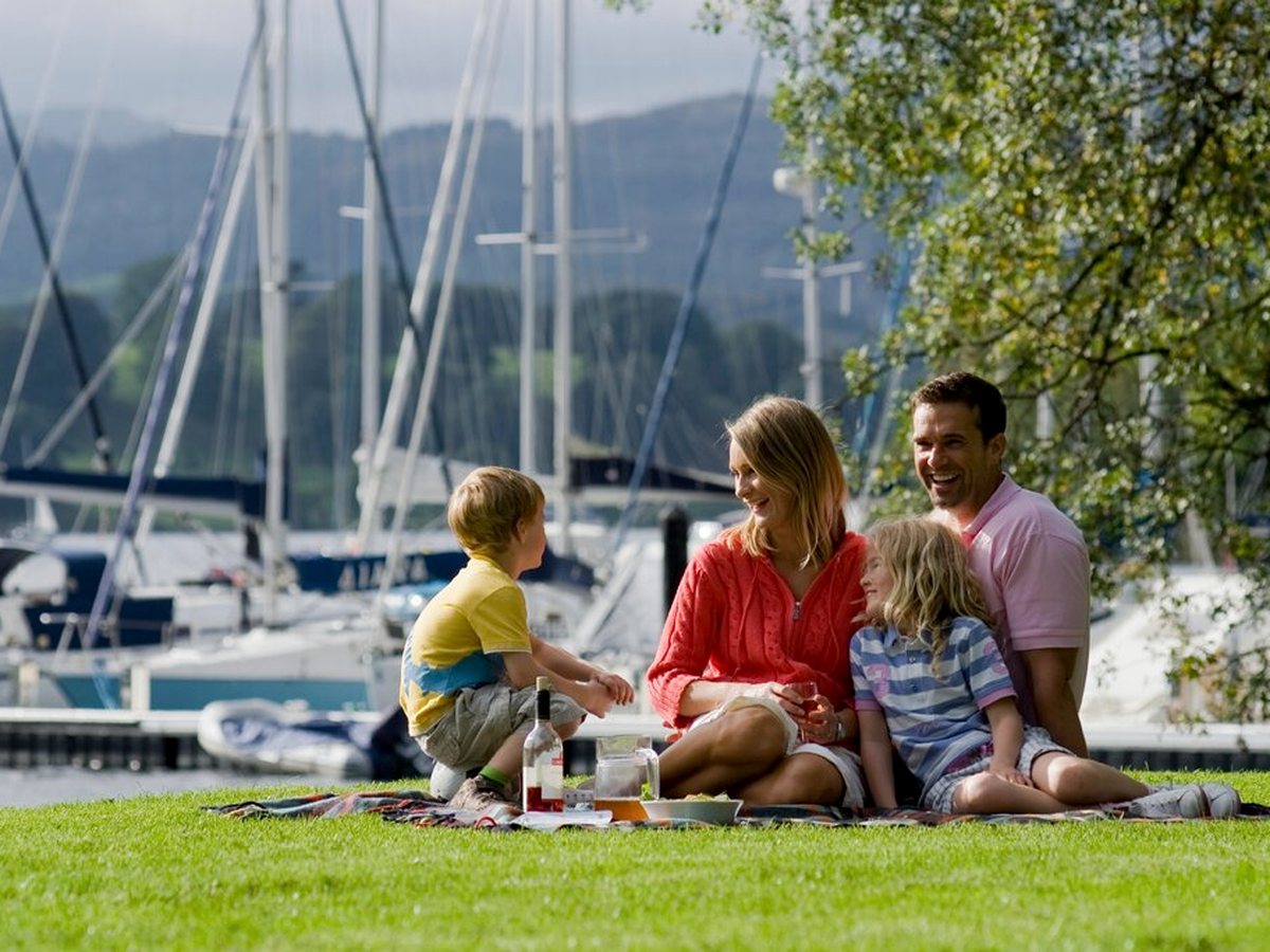 A family of four sitting on a picnic blanket on a grassy lakeside lawn, with the masts of moored yachts along the marina behind them and wooded hills across the water.