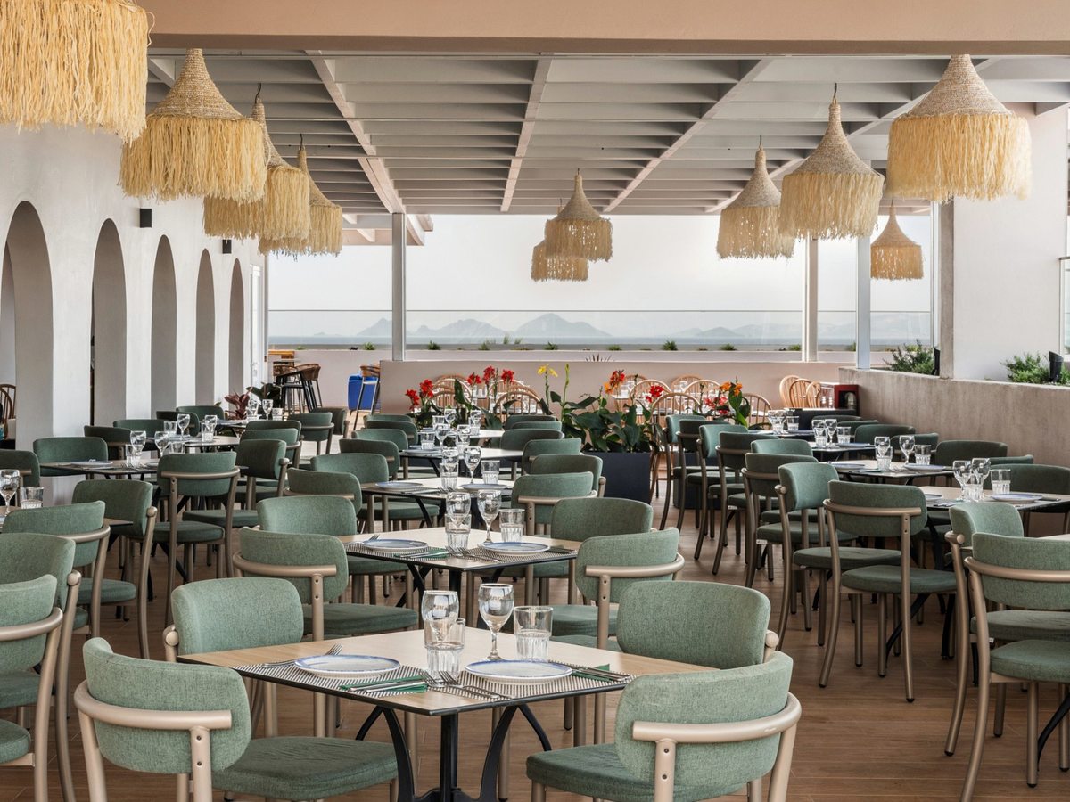 Covered open-sided dining terrace with green upholstered chairs, laid tables, raffia pendant lights and planted flowers, looking out over the sea to distant hills.