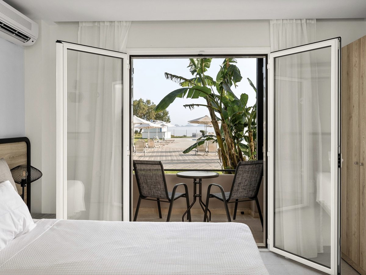 Double bed beside open glazed doors onto a private terrace with two chairs and a table, with banana plants, sun loungers and the sea beyond.