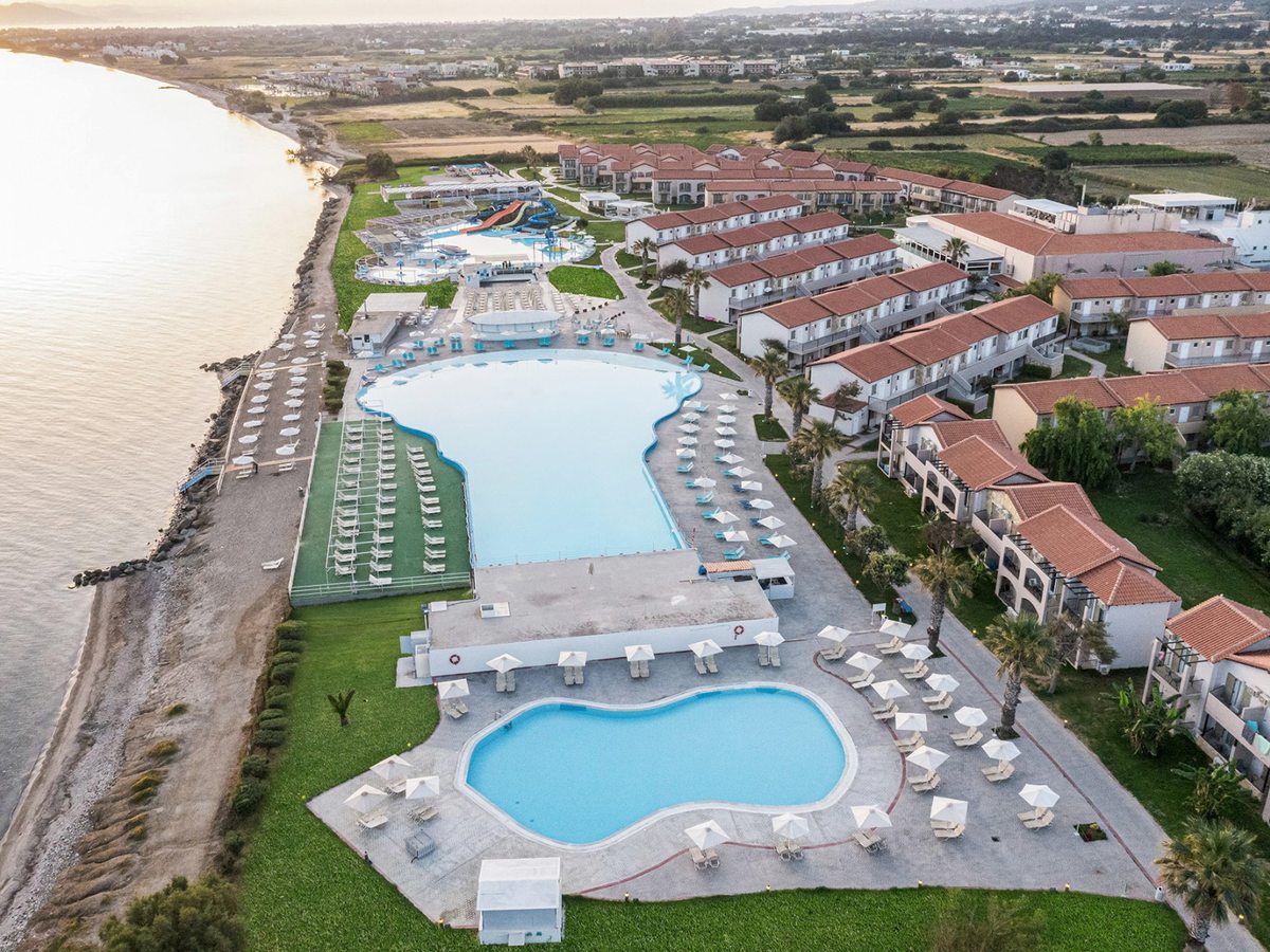 Aerial view of the shoreline: a narrow band of shingle and rock at the water's edge with rows of white sun loungers and parasols above it, and the large pool and room blocks behind.