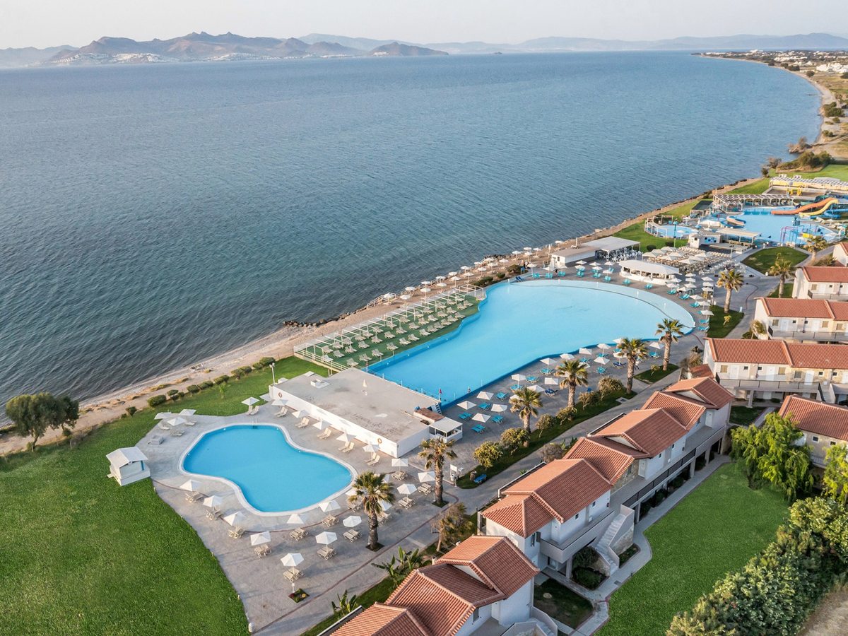 Aerial view along a wide bay, with the resort's pools and red-roofed buildings on the shore and the coastline curving away towards hills across the water.