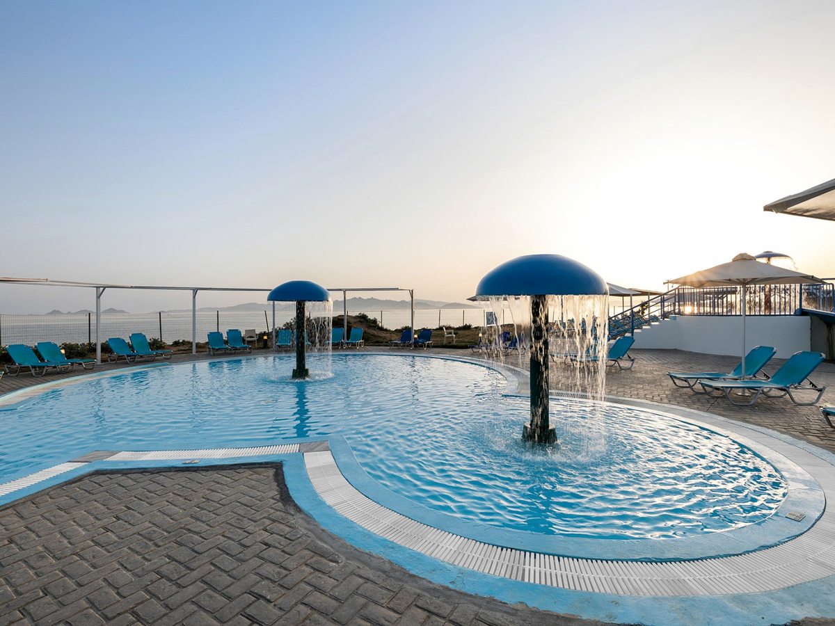 Small rounded shallow pool with two blue mushroom fountains pouring water, ringed by paving, blue sun loungers and parasols, with the sea beyond.
