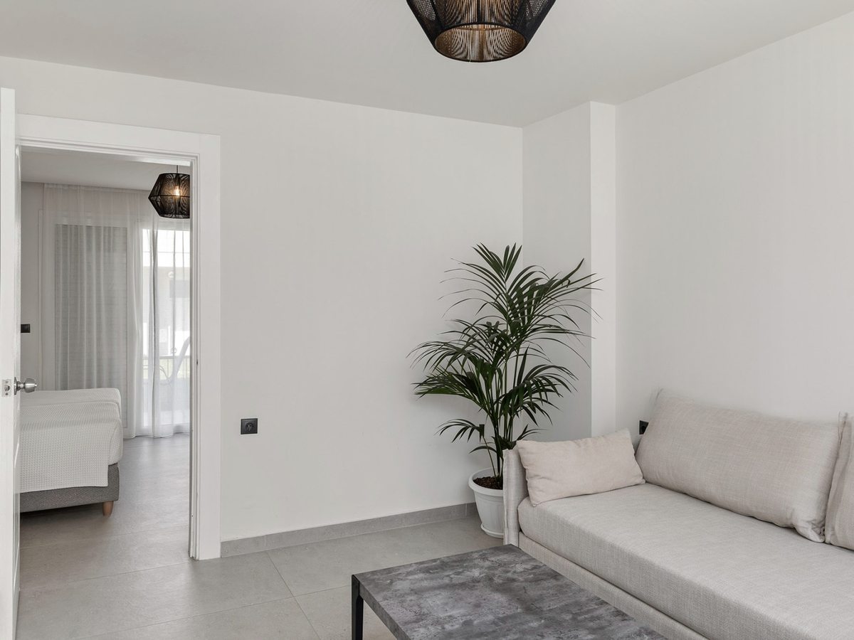 Pale sofa and a low coffee table in a white-walled sitting room with a potted palm and a woven pendant light, and an open door through to a bedroom.