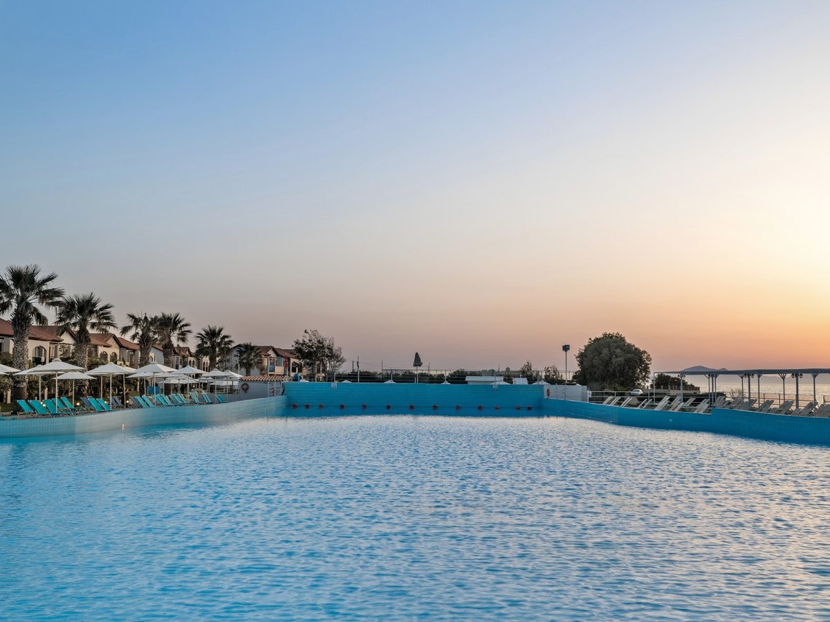 Very large blue pool with a tiled wall along its far side, sun loungers and white parasols on the terrace beyond, palm trees and red-roofed room blocks to the left and the sea to the right.